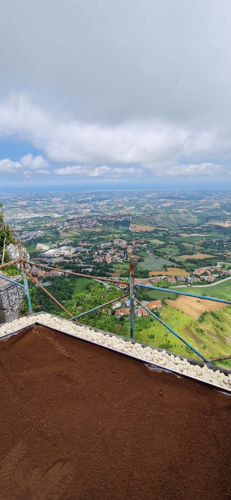 Aerial view of rolling countryside with patchwork fields and a blue river, seen from a high vantage point with railing and reddish-brown ground in foreground