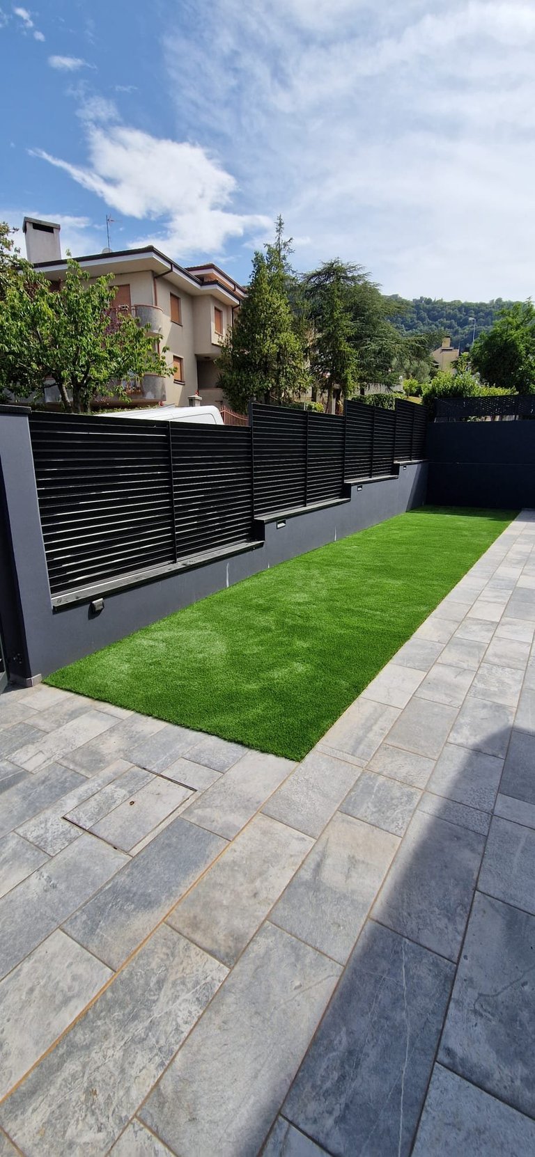 Modern backyard with manicured green lawn, black horizontal slat fence, stone patio, and two-story house in background under blue sky
