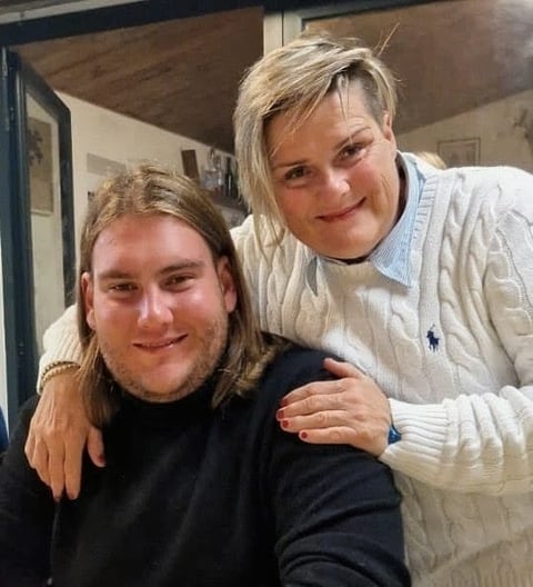 Two people smiling at the camera indoors, with one seated and one standing behind with arm around their shoulder