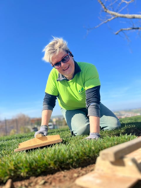 Woman in neon green shirt and sunglasses crouching on grass with landscaping tool on a clear day