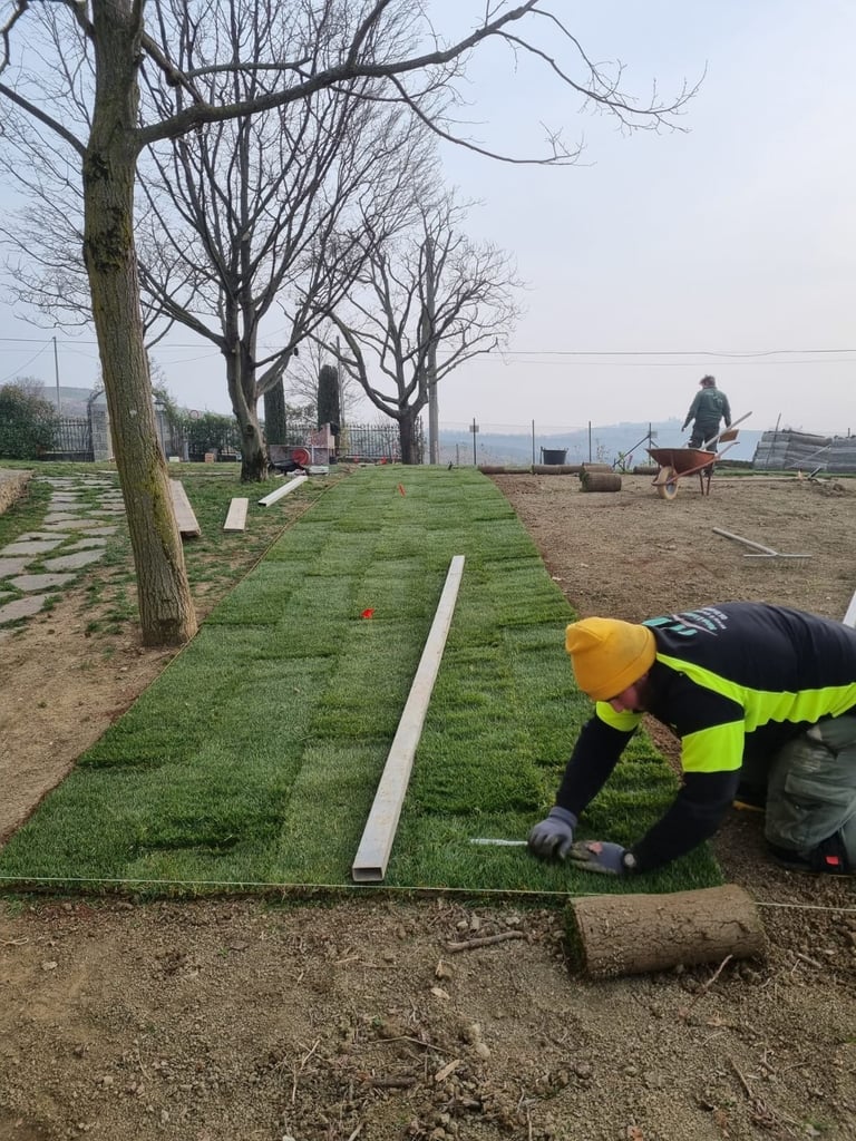 Worker in yellow hard hat and safety vest laying sod in grassy area with bare trees and other workers visible in background