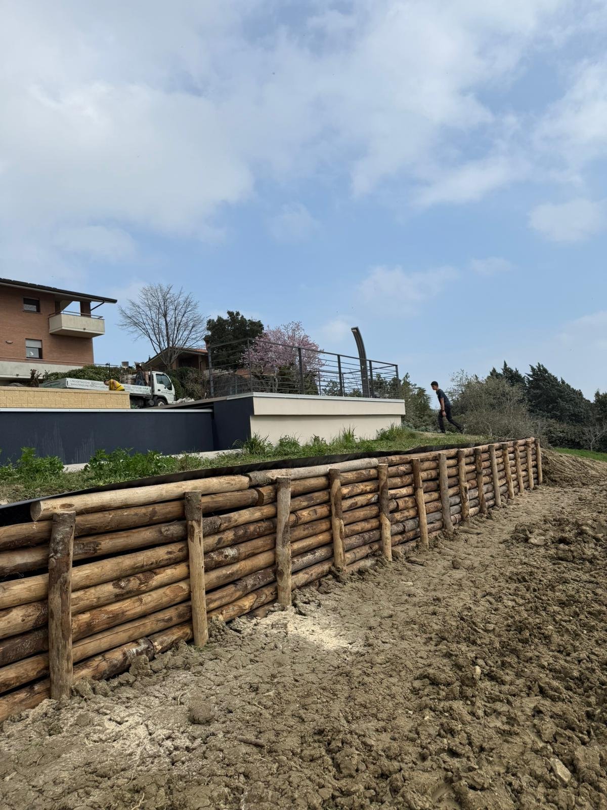 Wooden log fence lining a dirt path beside residential buildings with blooming trees and blue sky
