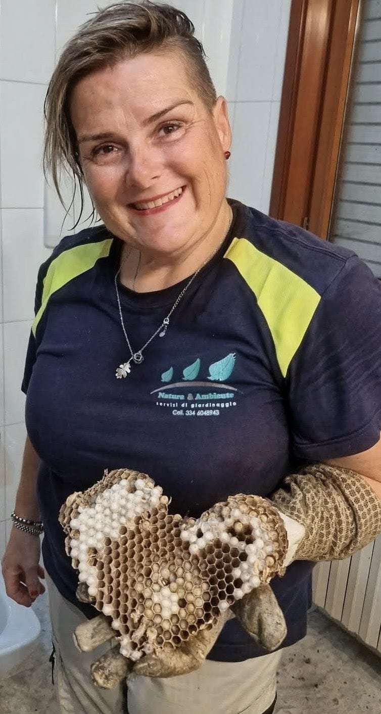 Woman holding a large wasp nest, smiling at camera while wearing protective gloves and a navy blue polo shirt