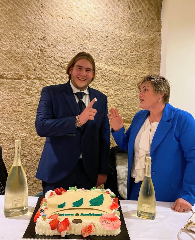 Two people in blue blazers stand at a table with a decorated cake and wine bottles, gesturing and celebrating indoors
