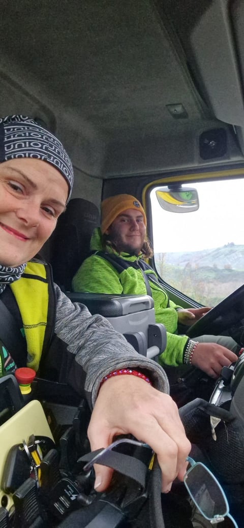 Two people in winter gear smiling inside a vehicle during a sunny mountain road trip